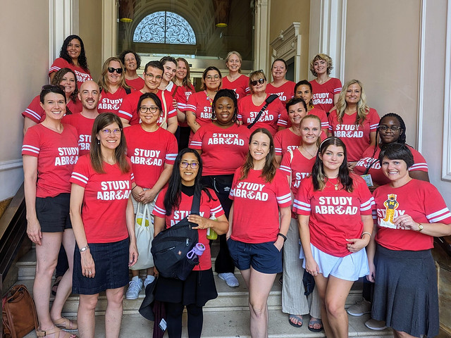 People wearing red shirts that stay "Study Abroad" pose on the steps of an old building.